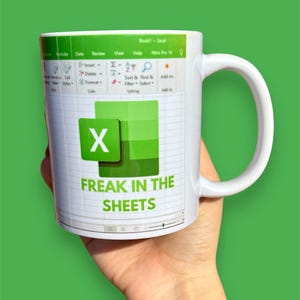 K&ouml;nnte beinhalten: Wei&szlig;e Keramiktasse mit einem gr&uuml;n-wei&szlig;en Design, das eine Excel-Tabelle mit dem Text "FREAK IN THE SHEETS" zeigt.