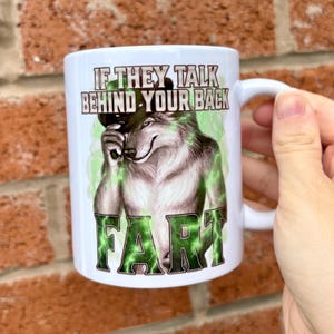 May include: A white ceramic mug featuring a graphic of a wolf wearing a hat, with the text "IF THEY TALK BEHIND YOUR BACK" above the wolf and the word "FART" in large green letters below, surrounded by a green glowing lightning effect.
