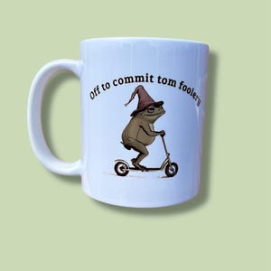 May include: White ceramic mug featuring a cartoon frog in a wizard hat riding a scooter. The mug is printed with the phrase "off to commit tom foolery" in a curved font.