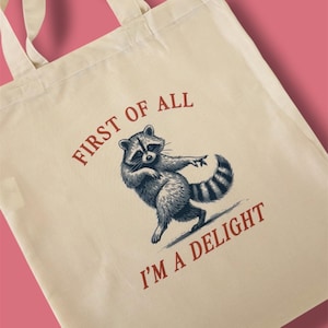 May include: A cream-coloured tote bag with a vintage-style illustration of a raccoon and the text "FIRST OF ALL I'M A DELIGHT" in red. The raccoon is depicted in black and white. The bag has two cream-coloured handles.
