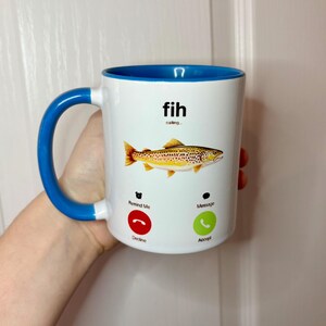 May include: White ceramic mug with a blue handle and interior. The mug features a fish illustration and the text "fih calling...". Below the fish are phone icons with the words "Remind Me" and "Message".