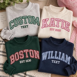 May include: Four crewneck sweatshirts in grey, cream, dark green, and navy blue. Each features embroidered collegiate-style lettering with a gingham pattern. The text reads CUSTOM TEXT, KATIE EST 2026, BOSTON EST 1630, and WILLIAM TEXT in various colours.