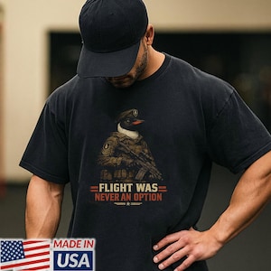 以下が含まれることがあります： 黒の半袖Tシャツ。軍用タクティカルギアを身にまといライフルを構えたペンギンのグラフィックが描かれています。グラフィックの下には、オレンジと白の文字で「FLIGHT WAS NEVER AN OPTION」と記され、小さな星のアイコンが添えられています。
