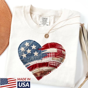 May include: Cream-colored t-shirt featuring a heart-shaped design resembling the American flag, with blue denim and white star accents. The shirt has a round neckline and a small gold necklace. The text "MADE IN USA" is visible.