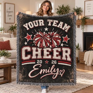 May include: A black woven throw blanket featuring a distressed texture, red and white pom-poms, and the text "YOUR TEAM CHEER 2026 Emily" with star accents. The blanket has a fringed border and a bold, athletic-style font design.