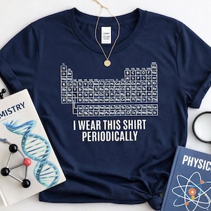 May include: Navy blue t-shirt with a white periodic table graphic and the text "I WEAR THIS SHIRT PERIODICALLY". The shirt is displayed with science books and a necklace. The design is suitable for science enthusiasts.