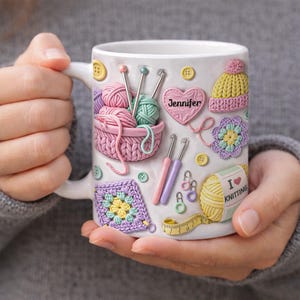 Könnte beinhalten: Weiße Keramiktasse, verziert mit 3D-Elementen zum Stricken und Häkeln. Mit einem Garnkorb, Häkelnadeln, einem Herz mit dem Namen "Jennifer" und dem Text "I love knitting".