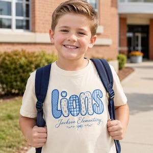 May include: A cream-coloured short-sleeve t-shirt featuring the word Lions in blue patterned bubble letters with a small heart above the i. Below the word, the text Jackson Elementary is printed in blue script with a small bow and leaf graphic.