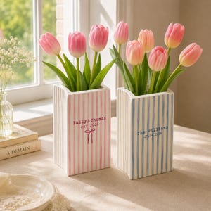 May include: Two rectangular ceramic vases with vertical stripes, one pink and one blue, each holding pink tulips. The pink vase features the text Emily & Thomas est. 2026 with a bow icon, and the blue vase displays The Williams 05.04.2026.