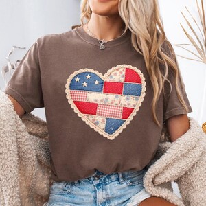 Peut inclure: Un t-shirt marron avec un motif en forme de cœur en patchwork. Le cœur présente des tissus rouges, blancs et bleus, avec une bordure en dentelle. Le motif comprend des étoiles, des motifs floraux et du vichy, évoquant un thème patriotique.