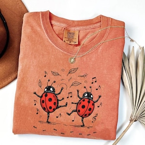 May include: A coral Comfort Colors t-shirt with a playful design of two dancing ladybirds with red spotted shells. The graphic includes musical notes and leaves. A gold necklace with a pendant is draped on the shirt.