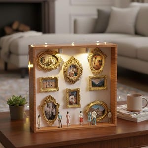 May include: A wooden shadow box with nine ornate gold-framed pictures of various sizes. The box is lit with small lights and contains miniature figures. The box is sitting on a wooden table with a mug and a plant.