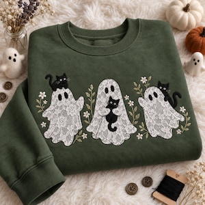 Könnte beinhalten: Ein olivgrüner Rundhals-Pullover mit einer Stickerei aus drei weißen Spitzengeistern. Jeder Geist wird von einer kleinen schwarzen Katze begleitet, wobei zarte Blumenstickereien und grüne Stängel zwischen den Figuren eingearbeitet sind.