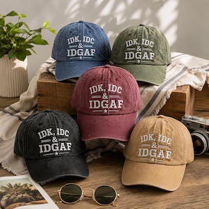 May include: Five baseball caps in blue, olive green, maroon, black, and tan, each featuring white embroidered text that reads IDK, IDC & IDGAF with horizontal lines and a small star. The caps have a distressed, washed-out fabric finish.