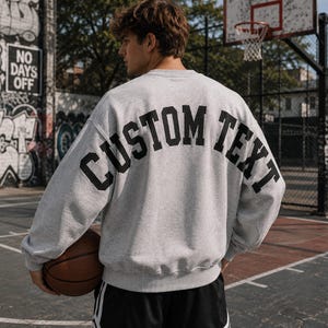 May include: A light gray crewneck sweatshirt featuring the black text CUSTOM TEXT printed in a large, arched varsity-style font across the upper back. The garment has long sleeves, a ribbed collar, and a ribbed hem, providing a relaxed fit.