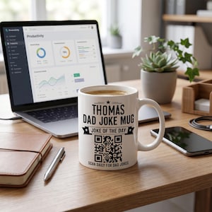 May include: A white ceramic mug featuring black text that reads Thomas Dad Joke Mug, Joke of the Day, and Scan Daily for Dad Jokes, along with a central QR code. The mug has a curved handle and is positioned on a wooden desk surface.