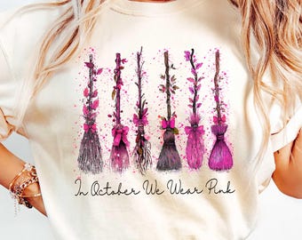 May include: Cream-colored t-shirt featuring watercolor-style illustrations of six witch brooms in varying shades of purple and pink. The brooms are adorned with bows and floral accents. The text "In October We Wear Pink" is printed below the brooms.