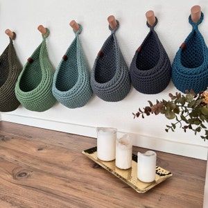 Puede incluir: Una fila de seis cestas de ganchillo hechas a mano, en forma de l&aacute;grima, en tonos verdes y azules, colgadas de clavijas de madera en una pared blanca. Cada cesta tiene un peque&ntilde;o detalle de cuero. Una bandeja dorada con velas descansa en el suelo de madera.