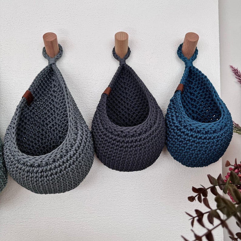 Puede incluir: Tres cestas colgantes de ganchillo en tonos grises y azules se exhiben contra una pared blanca. Cada cesta est&aacute; suspendida de una clavija de madera. Las cestas tienen forma de l&aacute;grima y est&aacute;n hechas de hilo grueso y texturizado. Una peque&ntilde;a etiqueta de cuero marr&oacute;n est&aacute; adherida al lado de dos cestas.