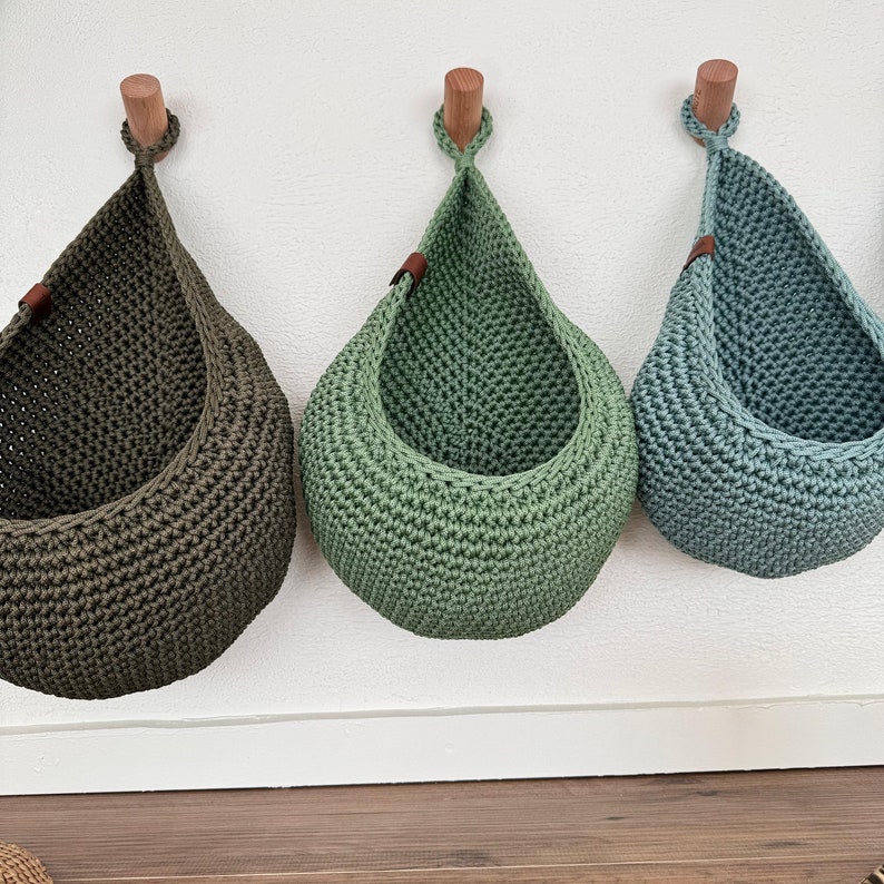 Puede incluir: Tres cestas colgantes de ganchillo en verde oliva, verde claro y verde azulado. Cada cesta, con forma de l&aacute;grima, est&aacute; suspendida de una clavija de madera con un detalle de cuero. Montadas en una pared blanca.