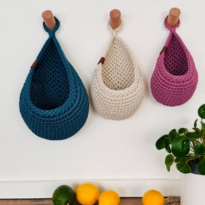 Puede incluir: Tres cestas colgantes de ganchillo en color turquesa, crema y malva, suspendidas de clavijas de madera. Las cestas en forma de l&aacute;grima est&aacute;n fijadas a la pared. Cada cesta tiene una peque&ntilde;a etiqueta de cuero. Las cestas est&aacute;n vac&iacute;as.