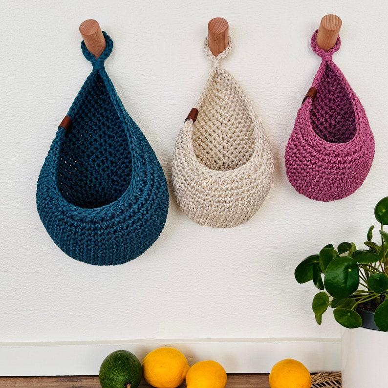 Puede incluir: Tres cestas colgantes de ganchillo en color turquesa, crema y malva, suspendidas de clavijas de madera. Las cestas en forma de l&aacute;grima est&aacute;n fijadas a la pared. Cada cesta tiene una peque&ntilde;a etiqueta de cuero. Las cestas est&aacute;n vac&iacute;as.