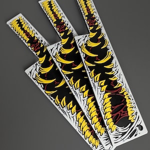 May include: Four white, rectangular stickers with a black, yellow, and red design. The design features a stylized, repeating pattern of yellow shapes against a black background, with red accents. The stickers are arranged on a gray surface.