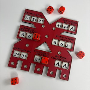 Puede incluir: Un tablero de juego rojo con texto y números blancos, y seis dados rojos. El tablero tiene secciones recortadas con texto como "Lethal Hits" y "Devastating Wounds". Los dados están esparcidos alrededor del tablero.