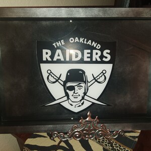 Peut inclure: Panneau encadré avec le logo des Raiders d'Oakland. Le logo est un bouclier noir et blanc avec le nom de l'équipe et un visage sur le thème des pirates avec des épées croisées. Le cadre est gris foncé avec une finition métallique.