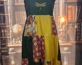 May include: A patchwork dress with a green bodice embroidered with a dragonfly. The dress features floral, polka dot, and solid fabric panels in yellow, red, and green. The straps are tied with floral fabric.