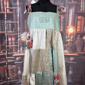 May include: A patchwork dress with a floral and quilted pattern. The dress features a square neckline, tie straps, and a tiered skirt. The bodice is a light green color with embroidered flowers. The dress is displayed on a black mannequin.