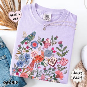 May include: A light purple t-shirt featuring a colorful graphic print of two birds surrounded by various flowers, leaves, and mahjong tiles. One tile displays the Chinese character for dragon. The shirt has a crew neckline and a Comfort Colors brand label.