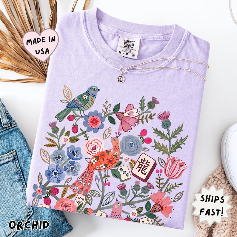 May include: A light purple t-shirt featuring a colorful graphic print of two birds surrounded by various flowers, leaves, and mahjong tiles. One tile displays the Chinese character for dragon. The shirt has a crew neckline and a Comfort Colors brand label.