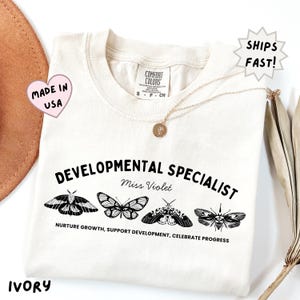 May include: An ivory t-shirt featuring black text that reads DEVELOPMENTAL SPECIALIST, Miss Violet, and NURTURE GROWTH, SUPPORT DEVELOPMENT, CELEBRATE PROGRESS. The design includes four detailed black and white illustrations of different moths.