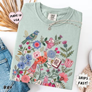 May include: A sage green t-shirt featuring a colorful graphic print of birds, various flowers, and mahjong tiles. The design includes a blue bird, a red bird, pink and blue blossoms, green foliage, and tiles with characters and dots. The shirt has a crew neck.