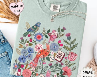 May include: A sage green t-shirt featuring a colorful graphic print of birds, various flowers, and mahjong tiles. The design includes a blue bird, a red bird, pink and blue blossoms, green foliage, and tiles with characters and dots. The shirt has a crew neck.