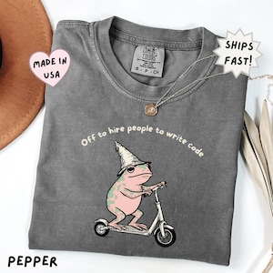 May include: A charcoal grey t-shirt featuring a graphic of a pink frog wearing a pointed hat and riding a scooter. The text above the frog reads, Off to hire people to write code. The shirt has a Comfort Colors brand label at the inner neckline.