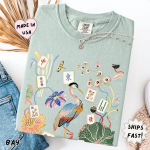 May include: A light sage green t-shirt featuring a colorful graphic print of a heron, lotus flowers, and various mahjong tiles. The shirt has a crew neckline and a Comfort Colors brand label at the inner collar. A silver chain necklace rests on top.
