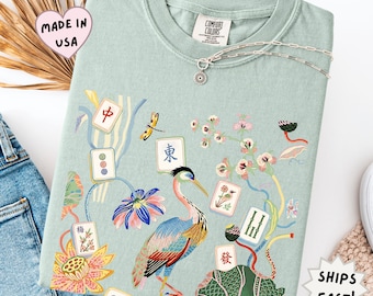 May include: A light sage green t-shirt featuring a colorful graphic print of a heron, lotus flowers, and various mahjong tiles. The shirt has a crew neckline and a Comfort Colors brand label at the inner collar. A silver chain necklace rests on top.