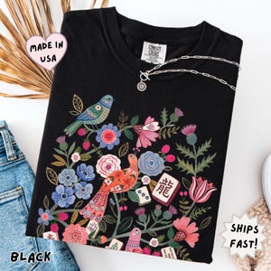 May include: A black crew neck t-shirt featuring a colorful graphic print of two birds surrounded by various flowers, foliage, and several mahjong tiles. The shirt has a Comfort Colors brand label at the inner neckline and a short-sleeved design.
