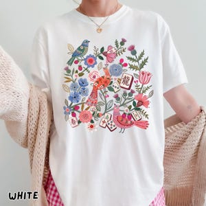 May include: A white short-sleeve t-shirt featuring a colorful graphic print of birds, flowers, and mahjong tiles. The design includes red, blue, and pink floral elements, green foliage, and several mahjong tiles with Chinese characters.