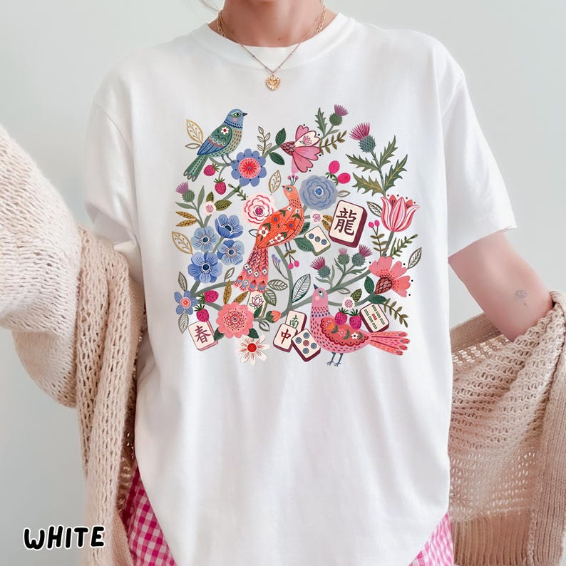May include: A white short-sleeve t-shirt featuring a colorful graphic print of birds, flowers, and mahjong tiles. The design includes red, blue, and pink floral elements, green foliage, and several mahjong tiles with Chinese characters.