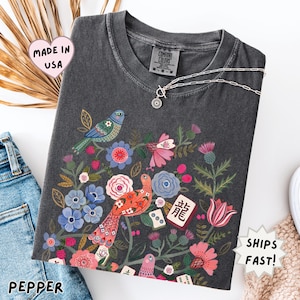May include: A charcoal gray short-sleeve t-shirt featuring a colorful graphic print of two birds surrounded by various flowers, foliage, and mahjong tiles. The shirt has a crew neckline and a Comfort Colors brand label on the inside collar.