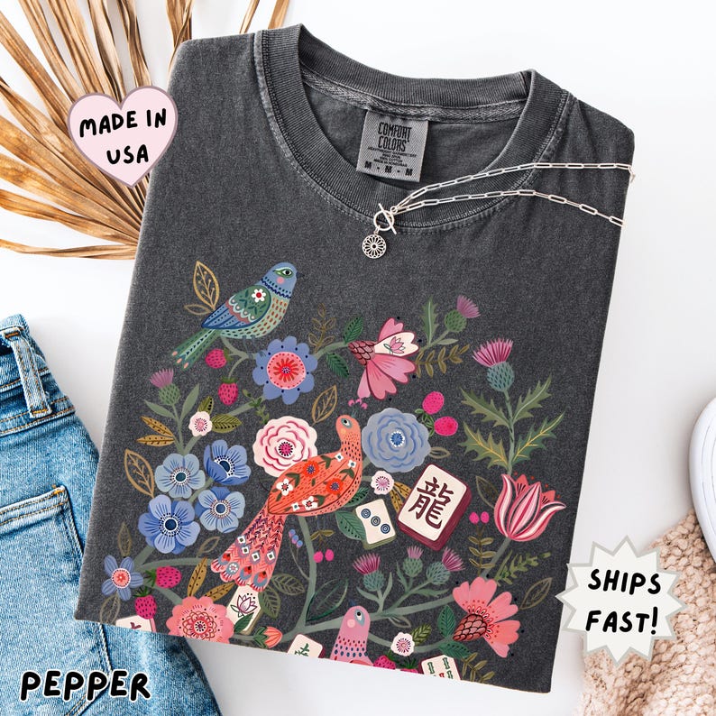 May include: A charcoal gray short-sleeve t-shirt featuring a colorful graphic print of two birds surrounded by various flowers, foliage, and mahjong tiles. The shirt has a crew neckline and a Comfort Colors brand label on the inside collar.