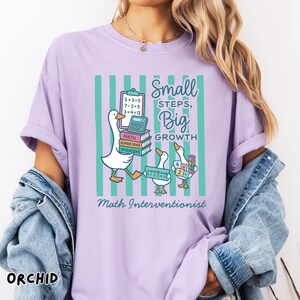 May include: A light purple t-shirt featuring a graphic of three white ducks with math tools, books, and a calculator. The text reads Small steps, Big growth and Math Interventionist over a background of vertical teal stripes on the shirt front.