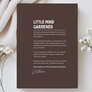 May include: A hardcover book with a dark brown cover. The title "LITTLE MIND GARDENER" is at the top, with text below describing the role. The name "Victoria" is signed at the bottom. White flowers are in the background.