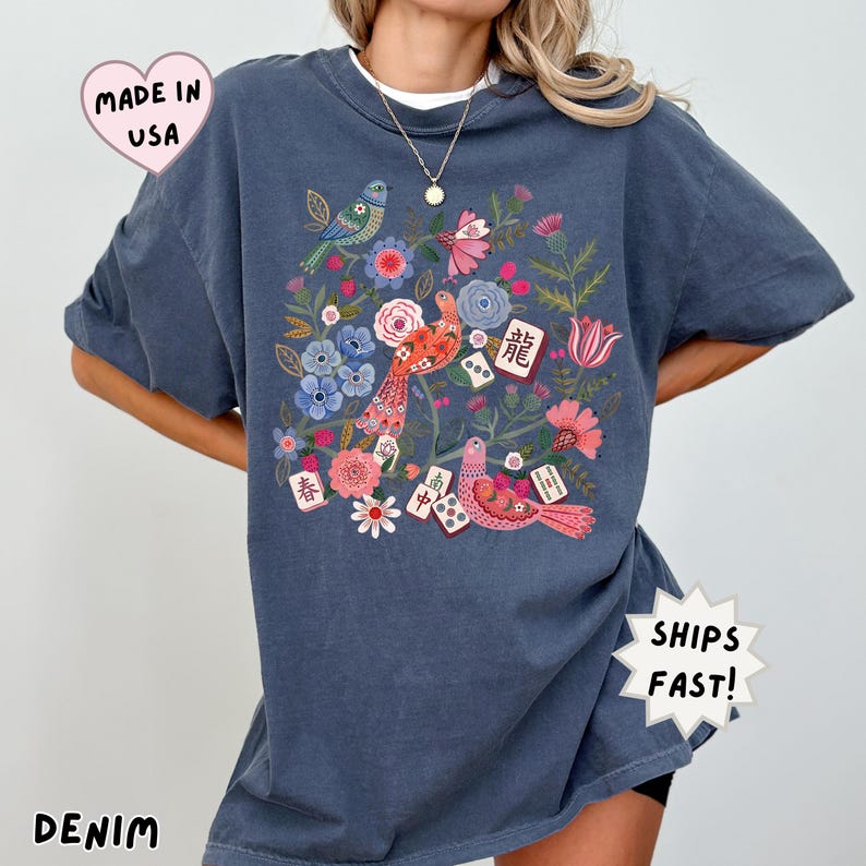 May include: A denim-colored t-shirt featuring a graphic print of colorful birds, various flowers, and mahjong tiles with Chinese characters. The design includes red, pink, blue, and green floral elements interspersed with game tiles.