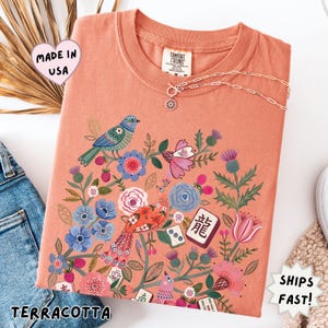 May include: A terracotta-colored t-shirt featuring a graphic print of a blue bird, a patterned peacock, various colorful flowers, and several mahjong tiles. The shirt has a crew neckline and a Comfort Colors brand label visible at the inner collar.
