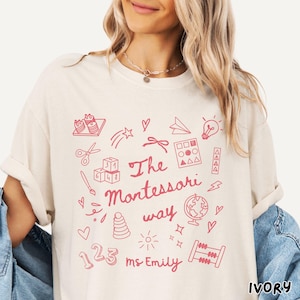 May include: Ivory-colored t-shirt with a pink doodle design. The design includes the text "The Montessori way ms. Emily" along with illustrations of a globe, abacus, and other educational symbols. The shirt is a relaxed fit.