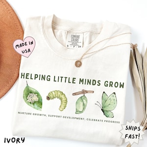 May include: Ivory-colored t-shirt with the text "Helping Little Minds Grow" and a watercolor illustration of a leaf, caterpillar, chrysalis, and butterfly. The shirt also has the text "Nurture Growth, Support Development, Celebrate Progress."